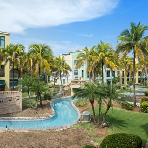 Escape to our tropical oasis with lush gardens and a meandering pool, framed by vibrant buildings and palm trees.