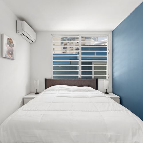 This white with blue themed bedroom is a perfect example of minimalist elegance. The cozy sleeping space is bathed in ample natural light, creating a serene ambiance that’s perfect for a restful stay.