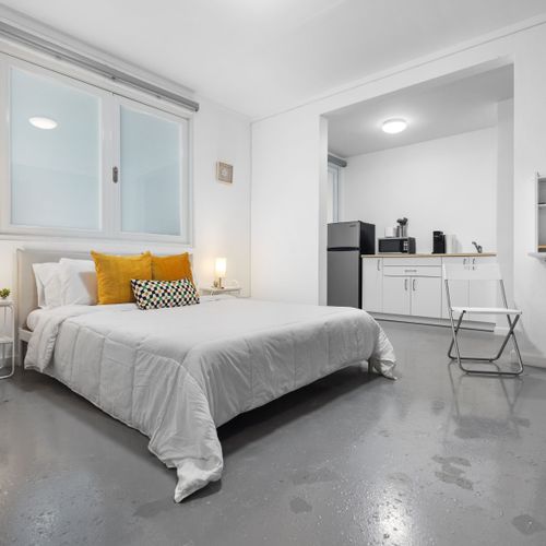 Inviting entry view of a modern studio apartment, showcasing a well-appointed bed with stylish bedding, conveniently placed lighting, and a peek into the simplistic kitchen space.