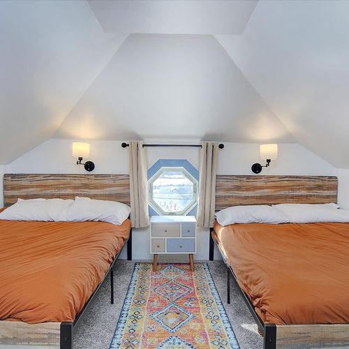 - inviting loft style bedroom with two full beds and warm, rustic headboards
- unique octagon window frames charming views and fills the space with light
- cozy carpet, soft lighting, and a colorful rug create a welcoming retreat