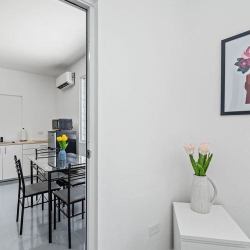 A sleek and airy dining area with a glass table, black chairs with slatted backs, and a vase of yellow flowers, featuring artworks of flowers, buildings, and abstract shapes on the walls.
