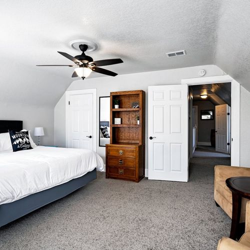 - spacious upper level bedroom with soft carpeting and a ceiling fan overhead
- features a plush bed, cozy armchair, and rustic wooden shelving for storage
- bright and clean with hallway access to additional guest spaces nearby