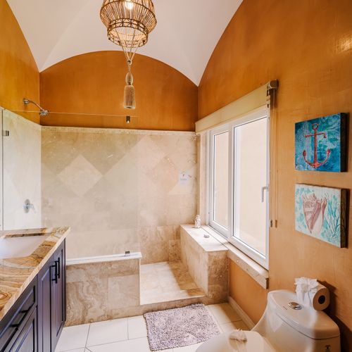 Master bath vertical view with rattan pendant, groin-vault ceiling, travertine walk-in shower, and tall window letting in natural light.