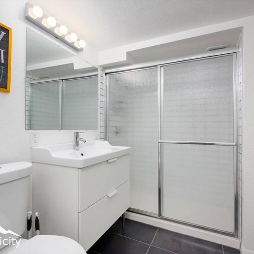 - sleek bathroom with a wide vanity, chrome faucet, and large mirrored light bar
- glass shower doors and black tile flooring add a polished, spa like vibe
- modern design meets comfort in this crisp and well lit space