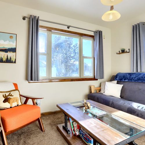 - bright and cozy living area featuring large windows that flood the space with natural light
- mid century orange accent chair with a moose pillow adds a fun lodge style charm
- rustic wood and glass coffee table perfect for games or morning coffee