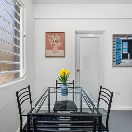 A bright and spacious dining area with a glass table, black metal chairs, and a vase of yellow flowers, surrounded by artworks of various styles and themes on the walls.