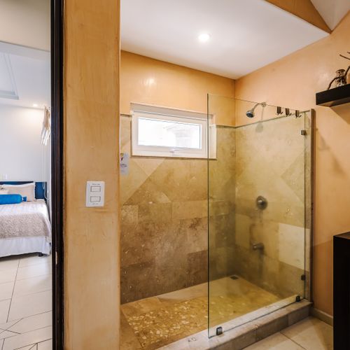 En-suite glass walk-in shower with travertine tile and view into the adjoining secondary bedroom with queen bed.