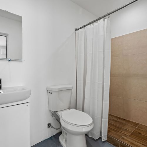 A white porcelain toilet sits adjacent to an equally white sink cabinet. Above the sink is a rectangular mirror mounted on the wall. The shower area is covered by a white curtain hanging from an overhead rod. Large beige tiles near the shower add an elegant touch, while wooden floor tiling provides warmth.