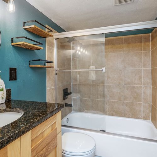- beautifully tiled tub shower combo with a sleek glass sliding door for a spa like feel
- granite countertop vanity with wood cabinetry and matte black fixtures
- wall mounted open shelves and round mirror add modern flair and functionality