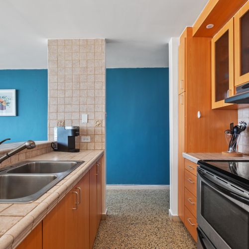 Fully equipped kitchen with double sink, tiled countertops, wooden cabinetry, and modern appliances including a stove and coffee maker.