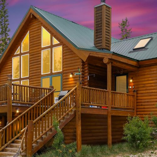 Rustic log cabin with warm interior lighting, spacious deck, and green metal roof glowing under the evening sky.