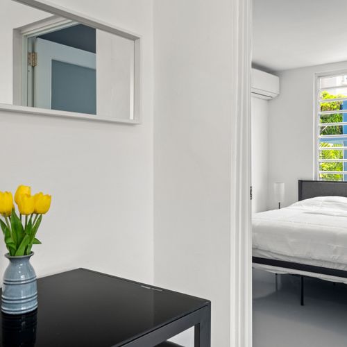 This modern bedroom is bright and airy, with ample natural light flooding in through the window. The walls are painted white, giving off a clean and minimalist vibe. A black table is prominent at the forefront, holding a vase with yellow tulips. Above the table on the wall is a rectangular mirror reflecting part of the room. There’s a bed to the right side covered in white bedding that contrasts nicely against the dark-toned bed frame. An air conditioning unit is mounted on the wall above the window.