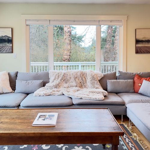 Bright and inviting living room with a spacious sectional sofa, cozy throws, and large windows overlooking peaceful woodland views.
