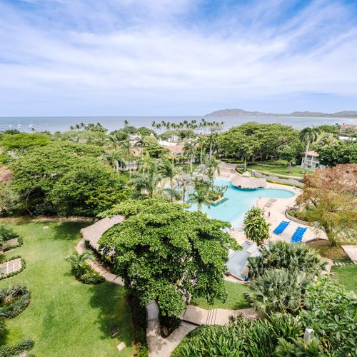 Aerial view of Matapalo Beach Resort lagoon pool, tropical gardens, and Tamarindo Bay beyond palm trees.