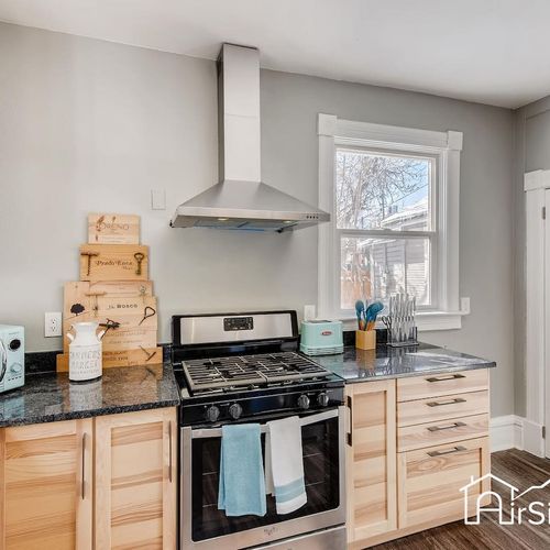 - bright kitchen featuring a gas range with vent hood, granite counters, and wood cabinetry
- decorative wine crates add a creative touch above the backsplash and window
- natural light fills the space for a welcoming and functional cook zone