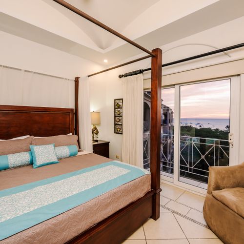 Master bedroom in morning light with four-poster king bed, wood ceiling fan, club chair, and ocean view through glass doors.