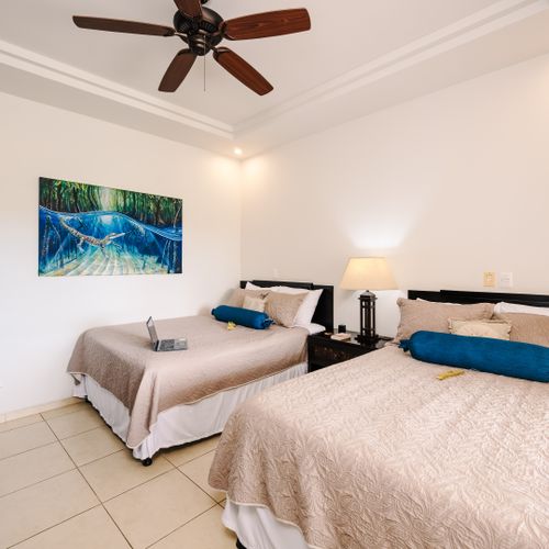 Guest bedroom with two beige-quilted beds, turquoise bolsters, wood ceiling fan, and tropical crocodile art.