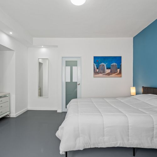 A minimalist bedroom with a white bed, a blue chair, and a beach painting.