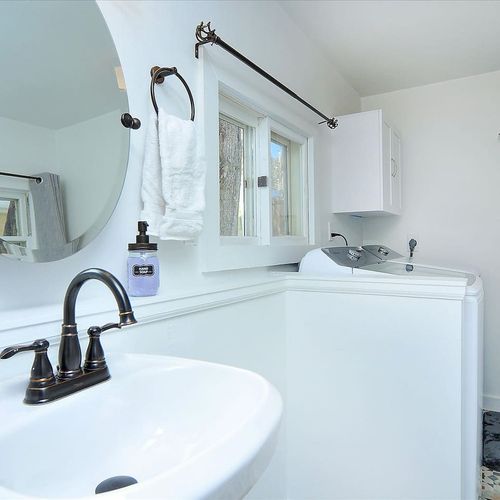- dual purpose space with a full size washer and dryer and modern pedestal sink
- sleek tile flooring, large round mirror, and updated fixtures throughout
- window views and clean layout make this room both practical and inviting
