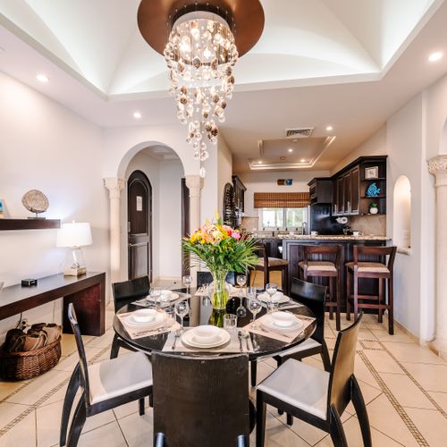 Dining room set for six under a crystal chandelier, flanked by arched columns, with kitchen breakfast bar visible through the archway.