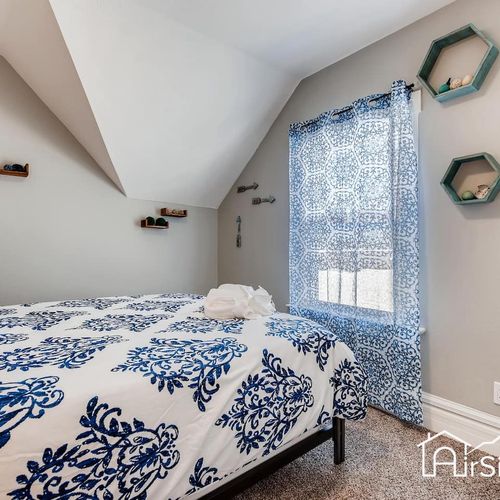 - soft blue drapes filter daylight into this peaceful room with cozy textures and neutral tones
- stylish wall shelving and angled ceiling add architectural interest and charm
- blue and white bedding brings a pop of pattern to the restful space