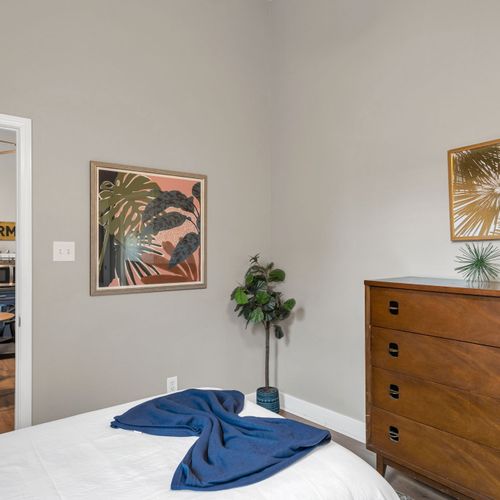 Sunlit corner w/ tropical artwork & a wood dresser — relaxed & full of character.