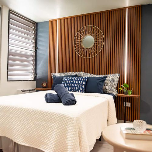 Discover comfort and style in this inviting bedroom. Featuring a warm wooden headboard with ambient lighting, a plush queen bed, and thoughtful blue accents, it's your perfect restful retreat.