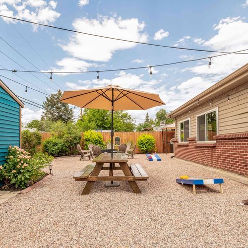 - bright and playful yard featuring a green doored blue shed and blooming garden beds
- central picnic table shaded by a large umbrella, ideal for outdoor meals
- string lights and fire pit circle invite cozy evenings under the stars