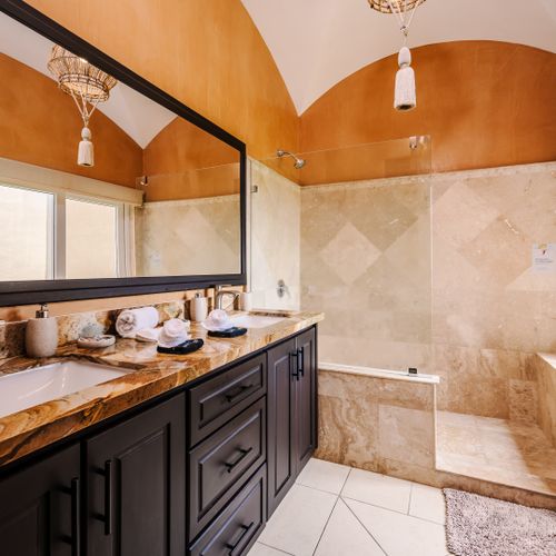 Master bathroom with dark-wood double vanity, marble countertop, large mirror, travertine walk-in shower, and ochre vaulted ceilings.