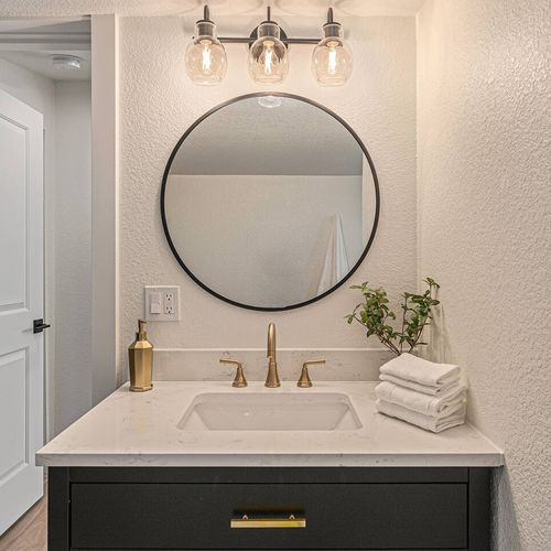 - stylish close up of the bathroom vanity highlights its round mirror, black cabinetry, & gold fixtures
- stacked towels & greenery add a fresh, welcoming feel to this clean & modern space
- perfect for getting ready in comfort with elegant touches