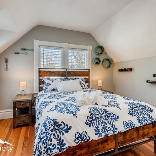 - bright attic style bedroom with rustic wooden bedframe and vibrant blue floral bedding
- matching nightstands with lamps sit beneath twin windows for perfect lighting
- wall accents and warm hardwood floors complete the cozy restful vibe