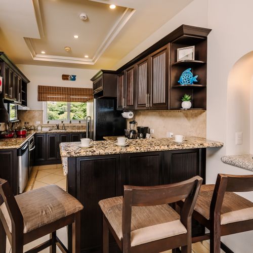 Kitchen breakfast bar with three upholstered stools, granite counter, dark cabinetry, round rattan mirror, and tropical wood sculpture.