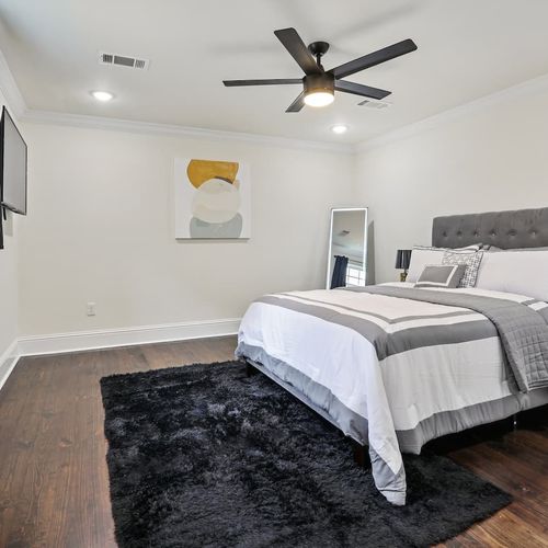 Clean, bright bedroom w/ a centered bed, dark rug & ceiling fan overhead.