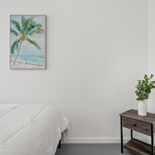 Relax in this cozy and bright bedroom by the sea, featuring a comfortable bed, a serene beach painting, and fresh greenery.