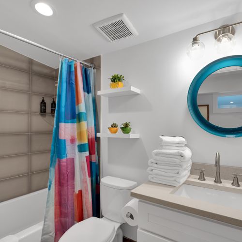 Bathroom 2 w/ a colorful shower curtain, teal mirror & neatly stacked fresh towels.