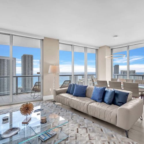 Spacious open-concept living space featuring a cozy sofa, dining area, and expansive windows showcasing stunning ocean and city views.