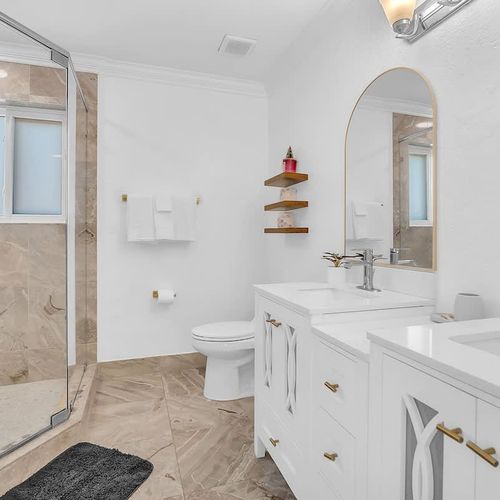 Bright, spa-like bathroom featuring a walk-in glass shower, dual vanities with elegant gold accents, and plenty of natural light.