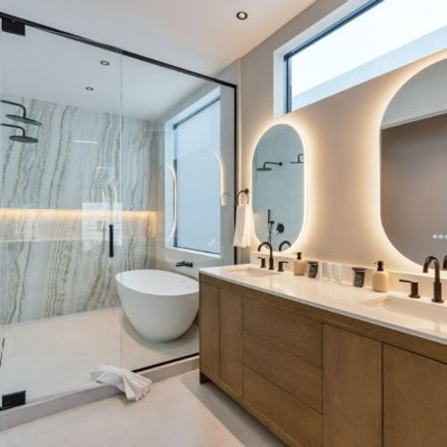Showcases sophisticated contemporary design and premium finishes throughout. Features include a sleek dual-sink vanity with ample storage, eye-catching oval backlit mirrors, and a striking freestanding bathtub situated under a bright privacy window.
