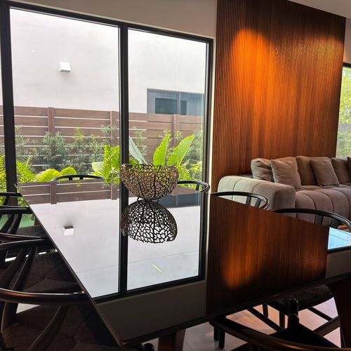 This sophisticated space highlights a seamless open-concept layout with rich wood wall paneling and floor-to-ceiling glass doors that invite natural light and views of the lush outdoor greenery.
