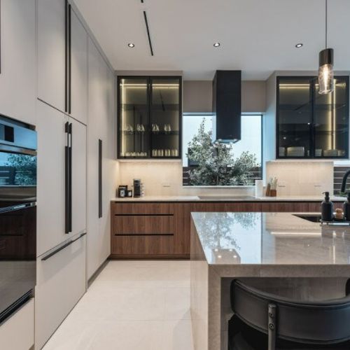 This high-end culinary space features a stunning contrast between warm wood lower cabinets and minimalist white floor-to-ceiling storage, anchored by a massive marble-style waterfall island.