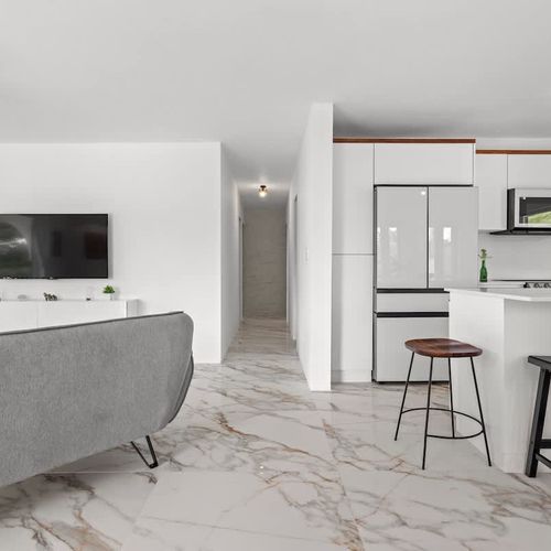 A spacious and modern open-plan living area with access to the balcony with views, with marble flooring, smart tv, ceiling fan, natural plants, coffee table and cozy grey sofa. lots of natural light and incredible views. white  kitchen with large cou