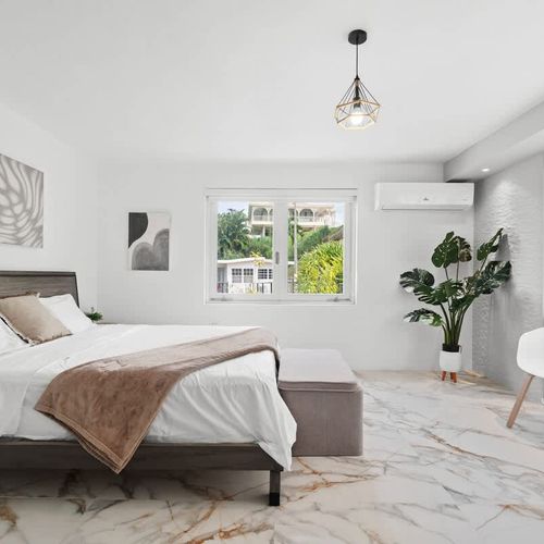 bright bedroom wiht king sized bed, lounge chair, wall art, ceiling lamp and windows and white curtains and air conditioner