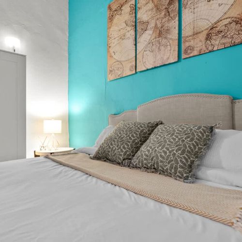 main bedroom with double bed, wall art, blue wall, large closet with mirror. bed side table with reading lamp and airconditioner. cushions and blankets