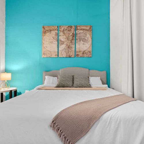 main bedroom with double bed, wall art, blue wall, large closet with mirror. bed side table with reading lamp and airconditioner. cushions and blankets