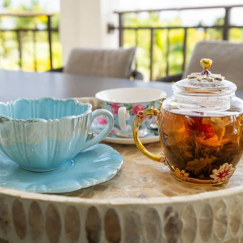 A charming outdoor patio features a tea set bathed in warm sunlight. A delicate blue teacup sits next to a glass teapot with vibrant tea, complemented by an elegant tray and a backdrop of lush greenery.