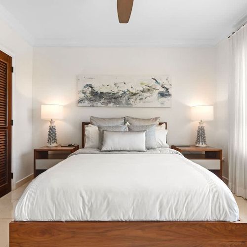 This serene bedroom provides a tranquil retreat with a plush bed in crisp white linens and a neutral palette. Natural light floods the room through large windows with stunning golf course views. A modern ceiling fan and air conditioning ensure comfor