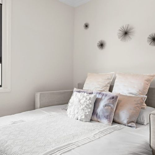 This inviting guest room features a comfy sofa bed with plush linens and decorative pillows. A neutral color palette and wood-paneled closet doors add warmth. Natural light creates a bright, airy feel, and a modern ceiling fan ensures comfort. A slee