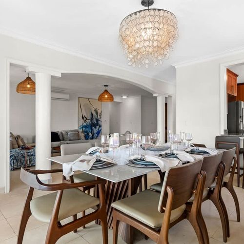 This elegant dining room oozes sophistication with its grand table, stunning chandelier, and neutral colors. Large windows offer beautiful views, and a sideboard and comfy sofa nearby add to the inviting atmosphere.