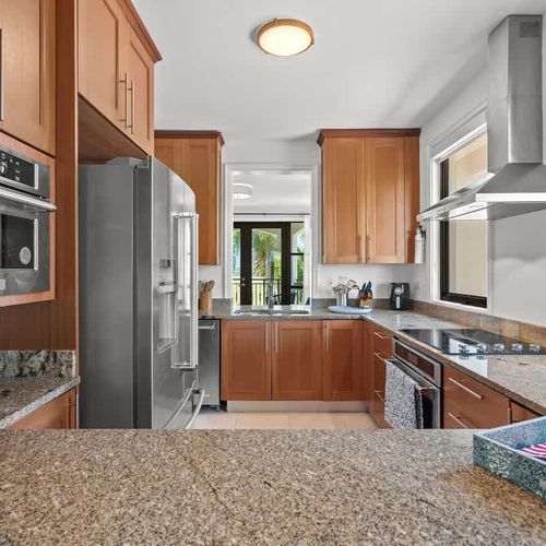 This modern kitchen has stainless steel appliances and warm wood cabinets. Granite countertops offer ample prep space, and the breakfast bar is perfect for casual dining. Large windows provide views of the lush tropical surroundings.