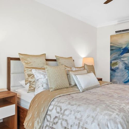 A spacious and elegant bedroom with a plush  bed dressed in luxurious linens and adorned with an array of throw pillows. The room features a neutral color palette with pops of gold, creating a serene and sophisticated ambiance.